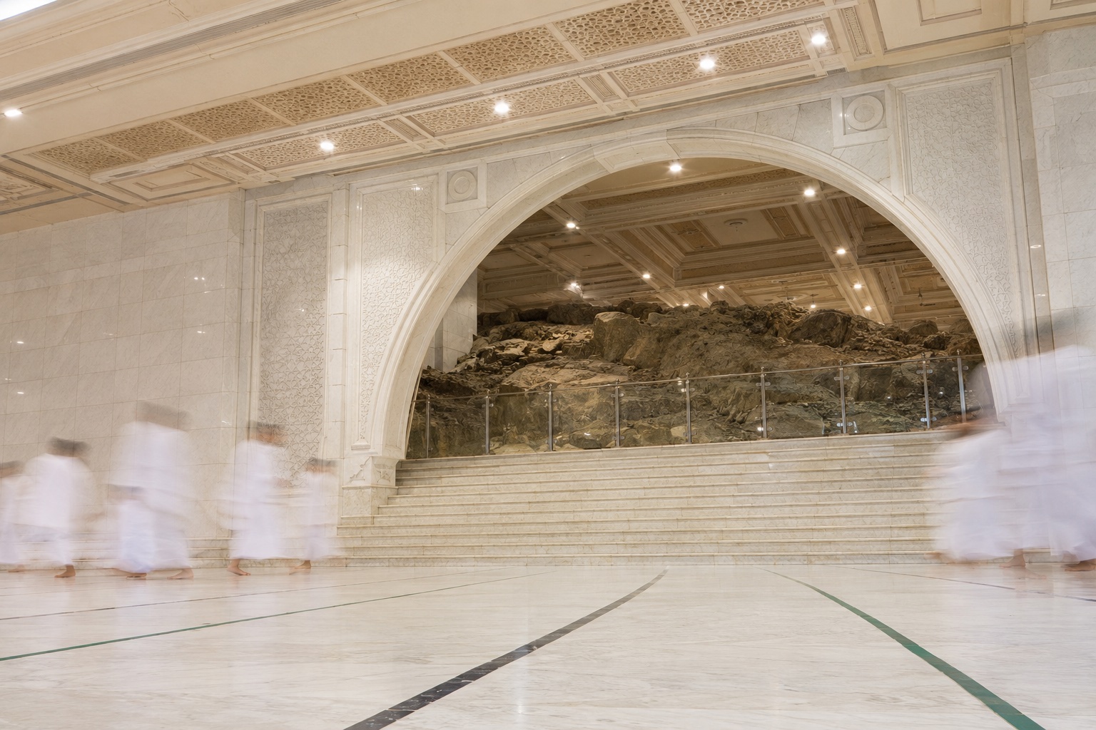 The marble steps and wide arch at Safa inside the mas'a gallery of Masjid al-Haram in Makkah, with the exposed rock of the hill behind railings and pilgrims walking past as soft motion blur