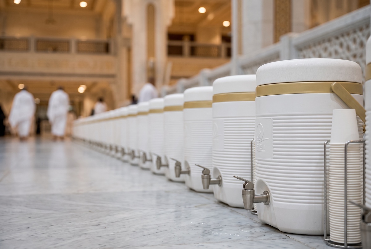 Rows of white and gold Zamzam water containers with taps and stacked cups standing on the polished marble floor of Masjid al-Haram in Makkah, pilgrims blurred in the background.