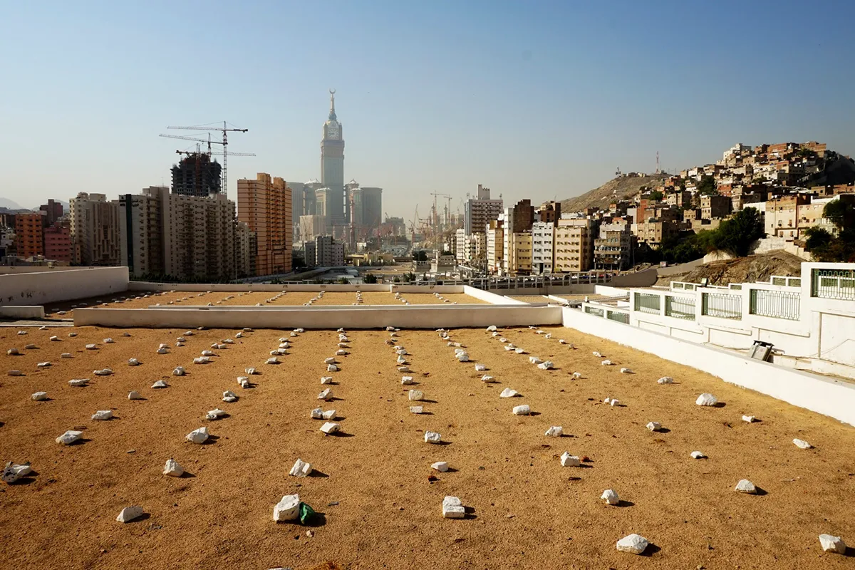 Rows of plain unmarked graves at Jannat al-Mu'alla cemetery in Makkah, bare stone and low boundary walls on the slope of al-Hajun, with the city rising behind in soft late-afternoon light.