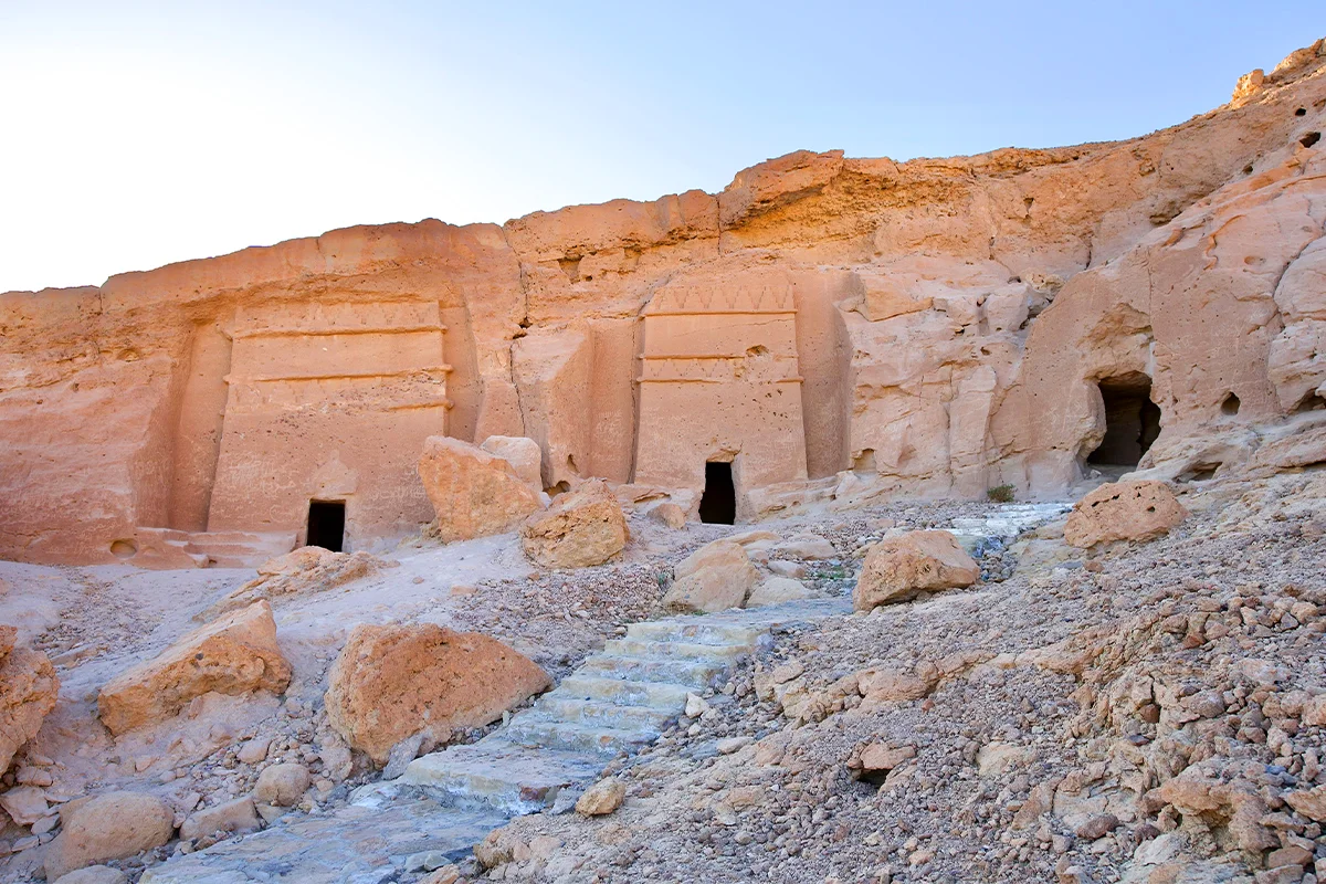 Low stone foundations and scattered pottery on open ground beside the palm oasis at al-Bad', with sandstone cliffs and rock-cut tombs behind.