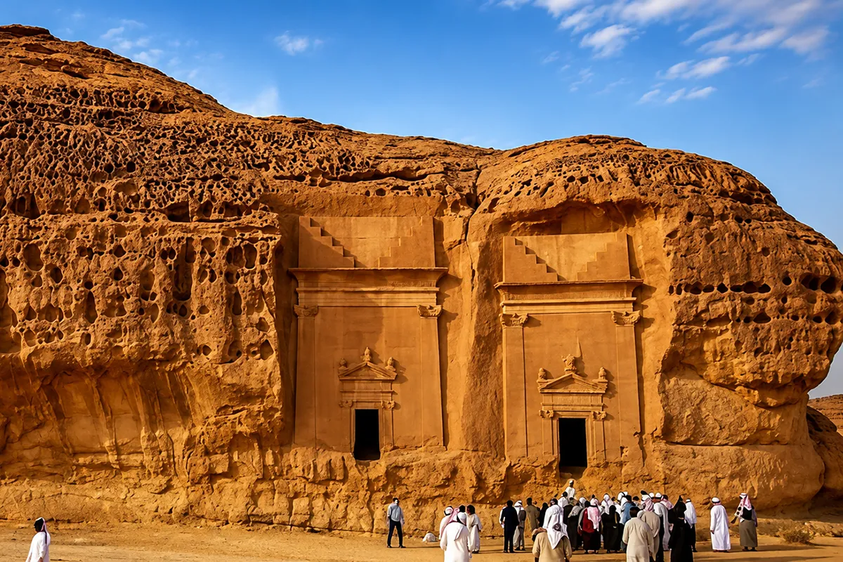 Square rock-cut tomb doorways with plain carved pillars in a weathered sandstone cliff near al-Bad', unrestored, in warm low light.