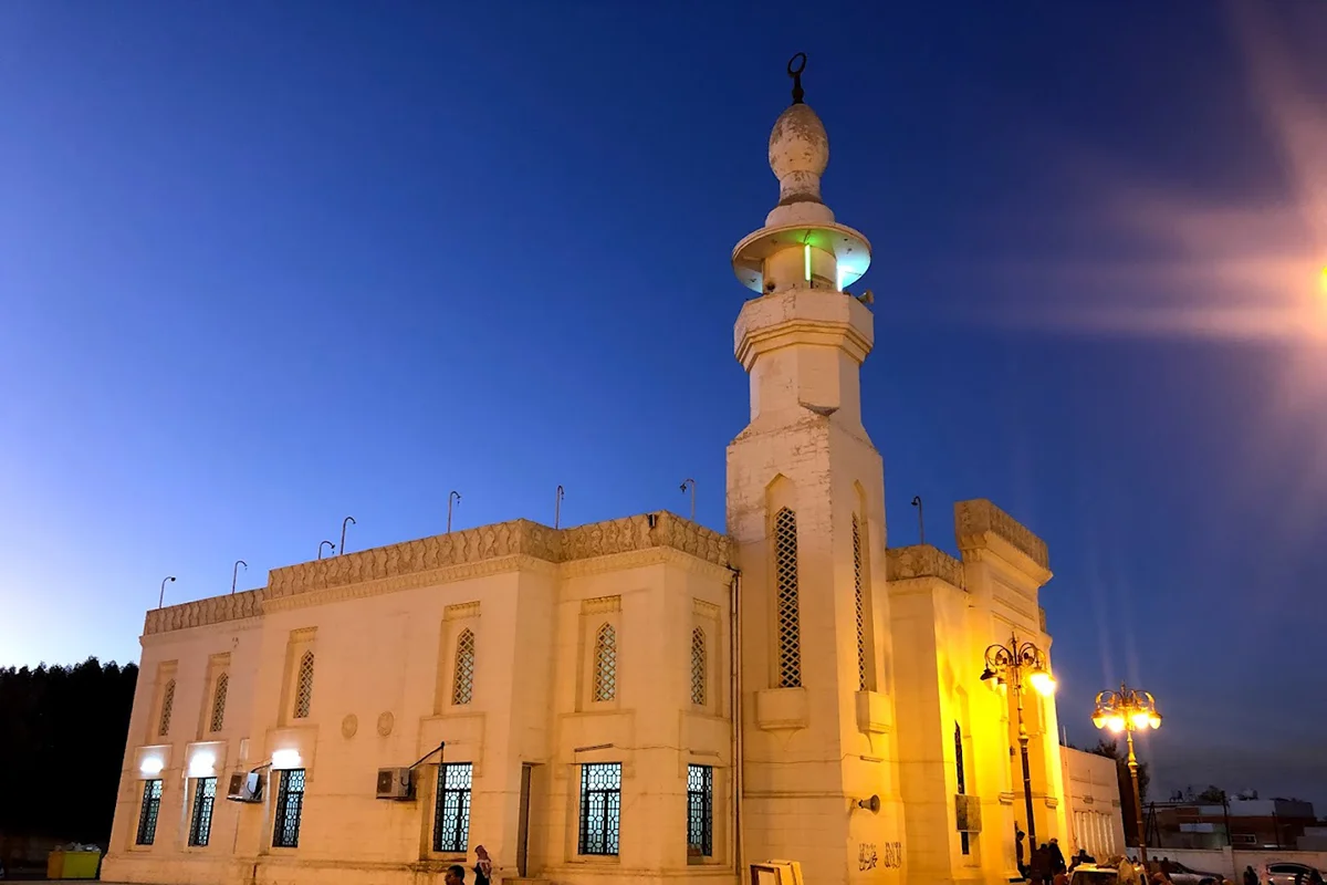 A plain modern mosque in Tabuk with a white facade, a single minaret and arched windows, seen from the street in clear daylight.