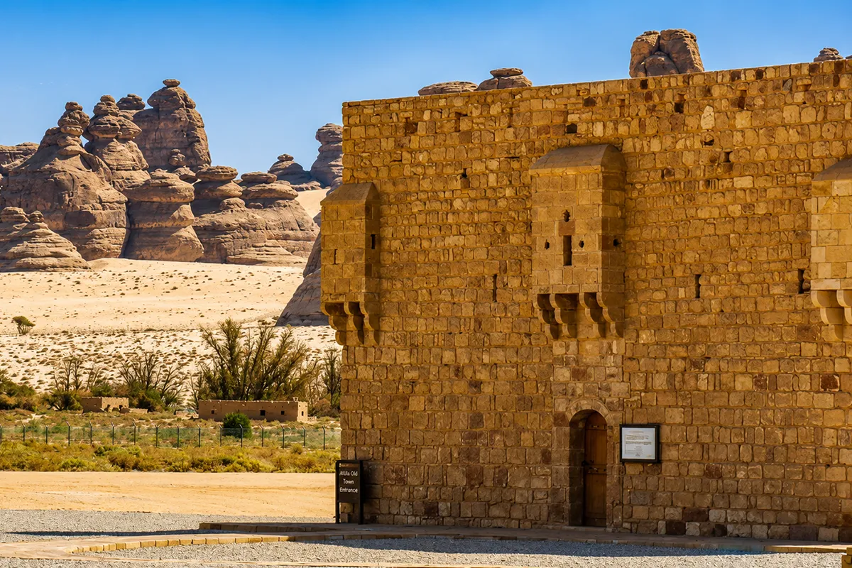 A square Ottoman stone fort with plain high walls and corner towers standing on open desert ground near Hegra, with sandstone outcrops behind it.