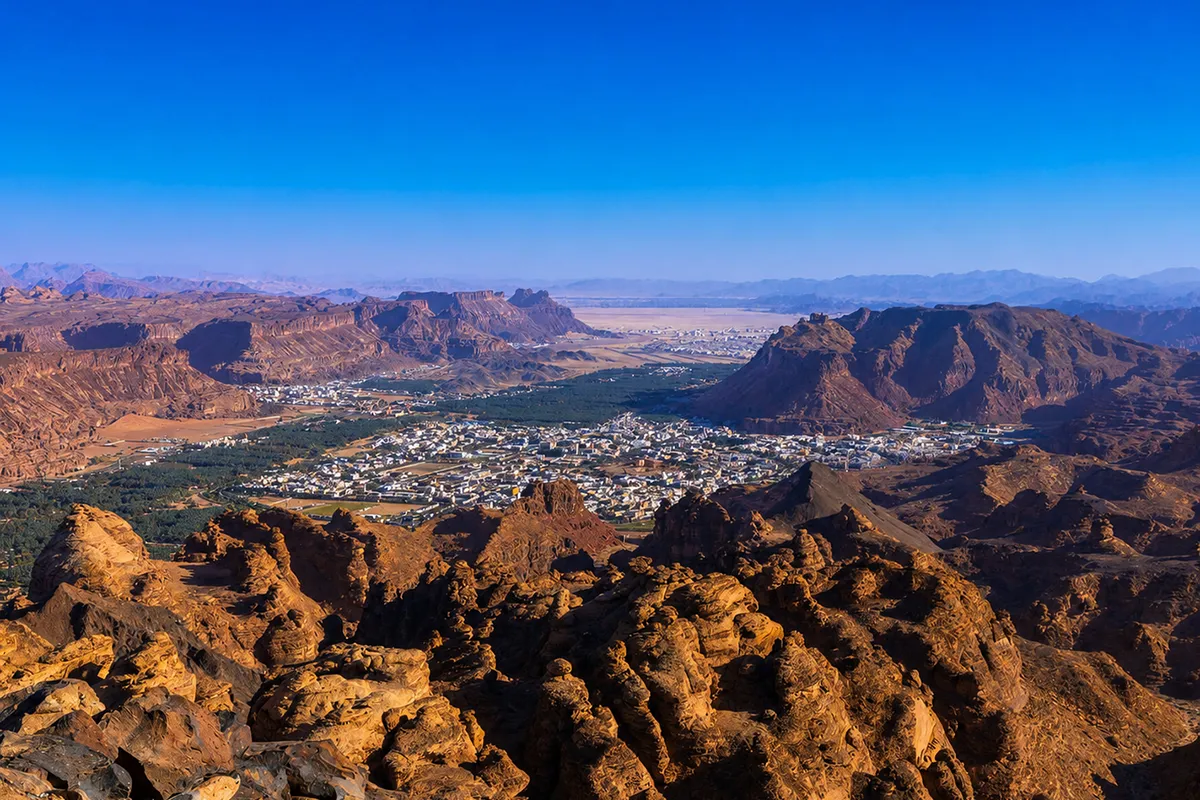 The AlUla valley floor with date palm plots and irrigation running between tall red sandstone cliffs, seen in warm late afternoon light.