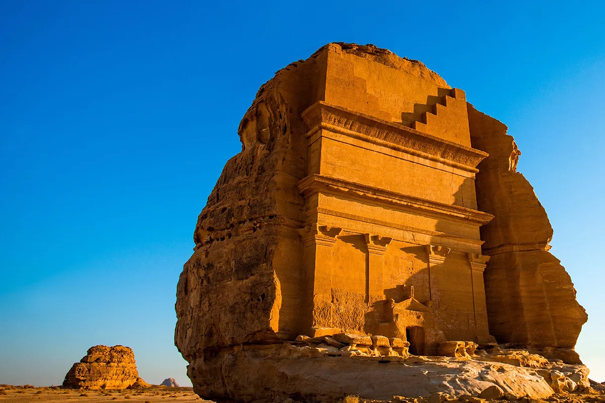 Bare red sandstone cliffs and open desert floor in the AlUla valley under hard daylight, with no buildings in view.