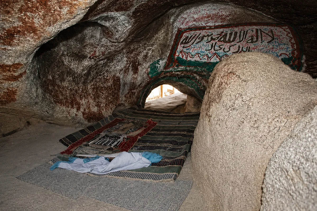 The low narrow western opening of Ghar Thawr among weathered boulders near the summit of Jabal Thawr, the interior in deep shadow, bare sunlit granite around it and the Makkah valley hazy far below.