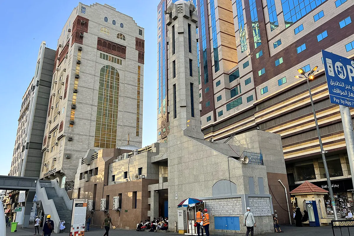 The pale stone facade and single minaret of Masjid al-Jinn in Makkah beside the Umar ibn al-Khattab ring road, with the wall of al-Mu'alla cemetery running along one side and Makkah's bare hills behind.