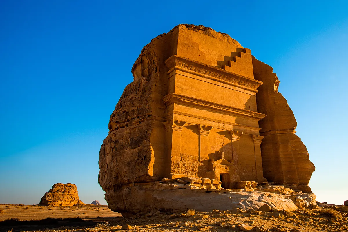 Monumental Nabataean tomb facades carved into isolated sandstone outcrops on the desert floor at Hegra in AlUla, weathered and unrestored, in low afternoon light.