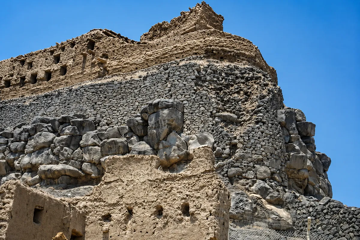 Ruined stone walls of a Khaybar stronghold on rocky ground, with other fortified outcrops visible in the distance across the oasis.