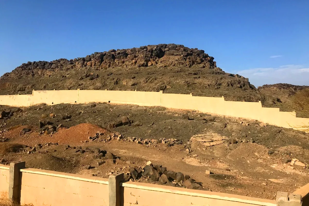 A low walled enclosure of plain unmarked graves on stony ground near the Khaybar oasis, with black lava country and palms in the distance.