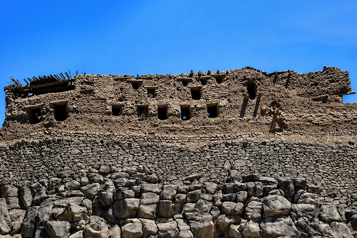 Broken stone fortress walls on a rocky rise above the Khaybar oasis, unrestored and weathered, with palms and black lava country behind.