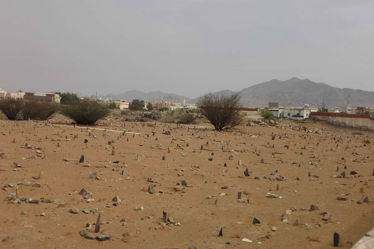 A low walled enclosure of plain unmarked graves on open gravel ground near Badr, with a metal fence and the empty valley beyond in morning light.