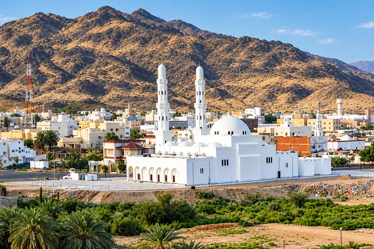 A small plain mosque standing on open gravel ground at the Badr battle site, with the wide valley floor and low ridges behind it in morning light.