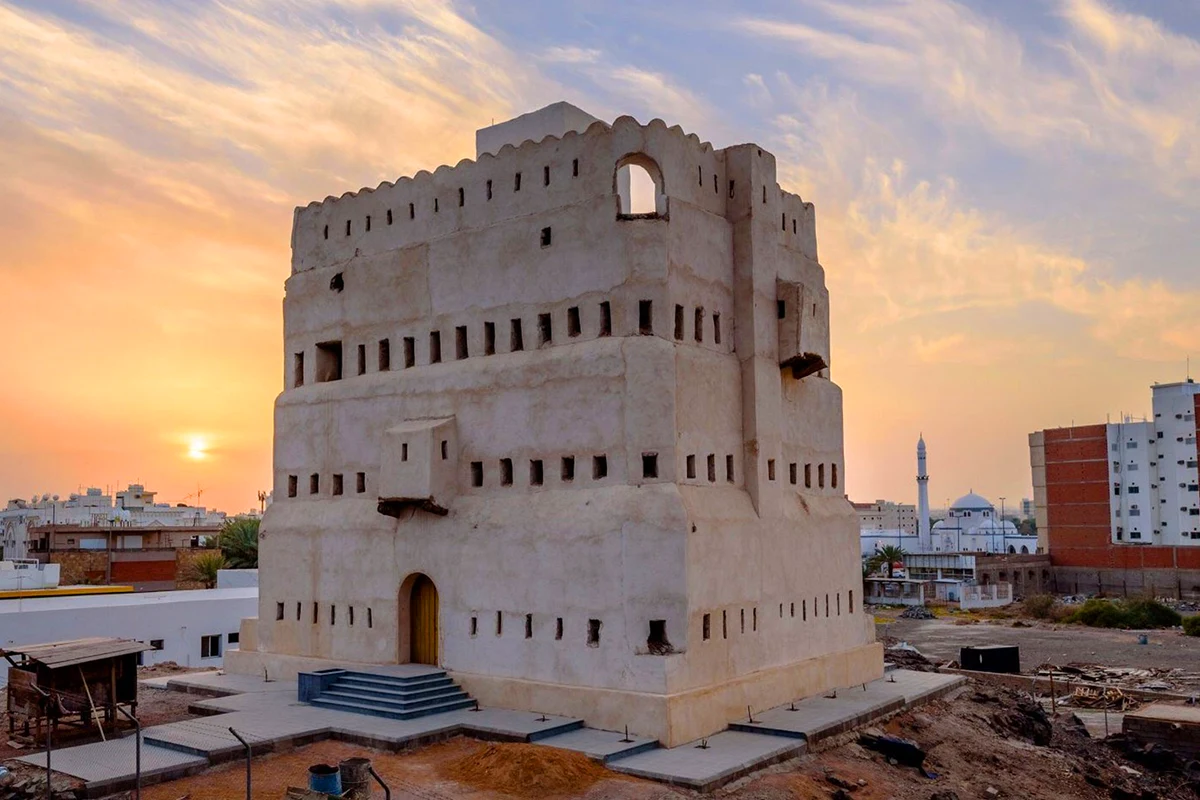 A compact three-storey stone Ottoman fort standing on a rocky plateau near Quba in Madinah, with small window openings and the city spreading below in afternoon light.