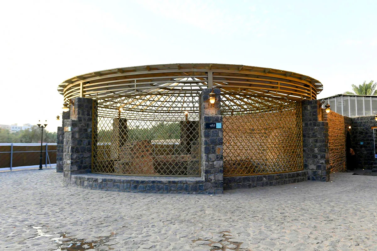 A stone-walled historic well enclosure with a staircase leading down to the water, surrounded by date palms in the Awali area of Madinah in morning light.
