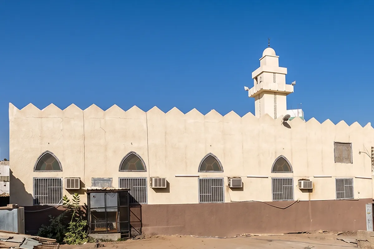 A small plain cream stone neighbourhood mosque with a low dome on a quiet Madinah street, seen in soft afternoon light with no people in view.