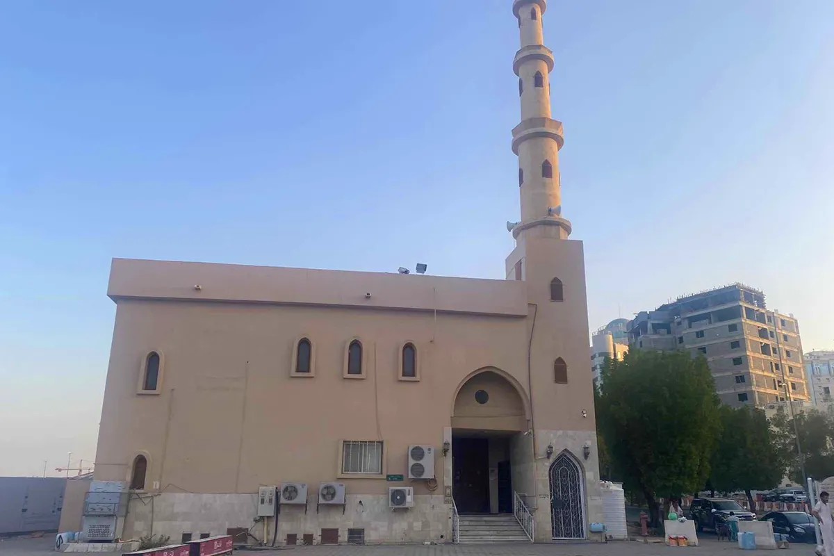 A very small plain cream stone mosque with a single slim corner minaret on a quiet street north of the Prophet's Mosque in Madinah, in warm afternoon light.