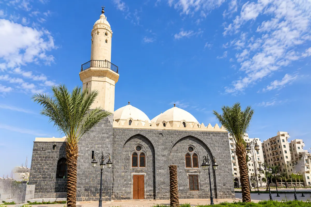 A quiet older quarter of Madinah with low walls, date palms and a narrow lane in warm afternoon light, on ground associated with an early mosque.