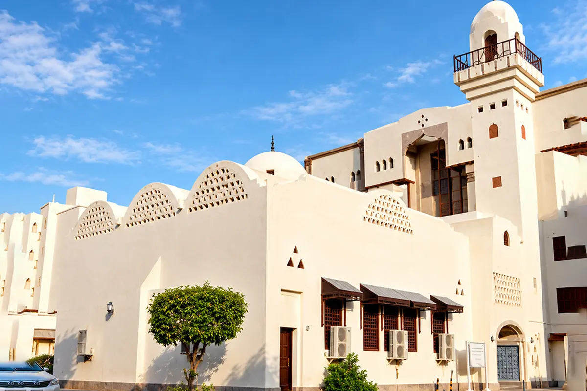 A small walled stone enclosure containing a plain historic mosque on the eastern side of Madinah, with modern buildings and the road beyond in afternoon light.