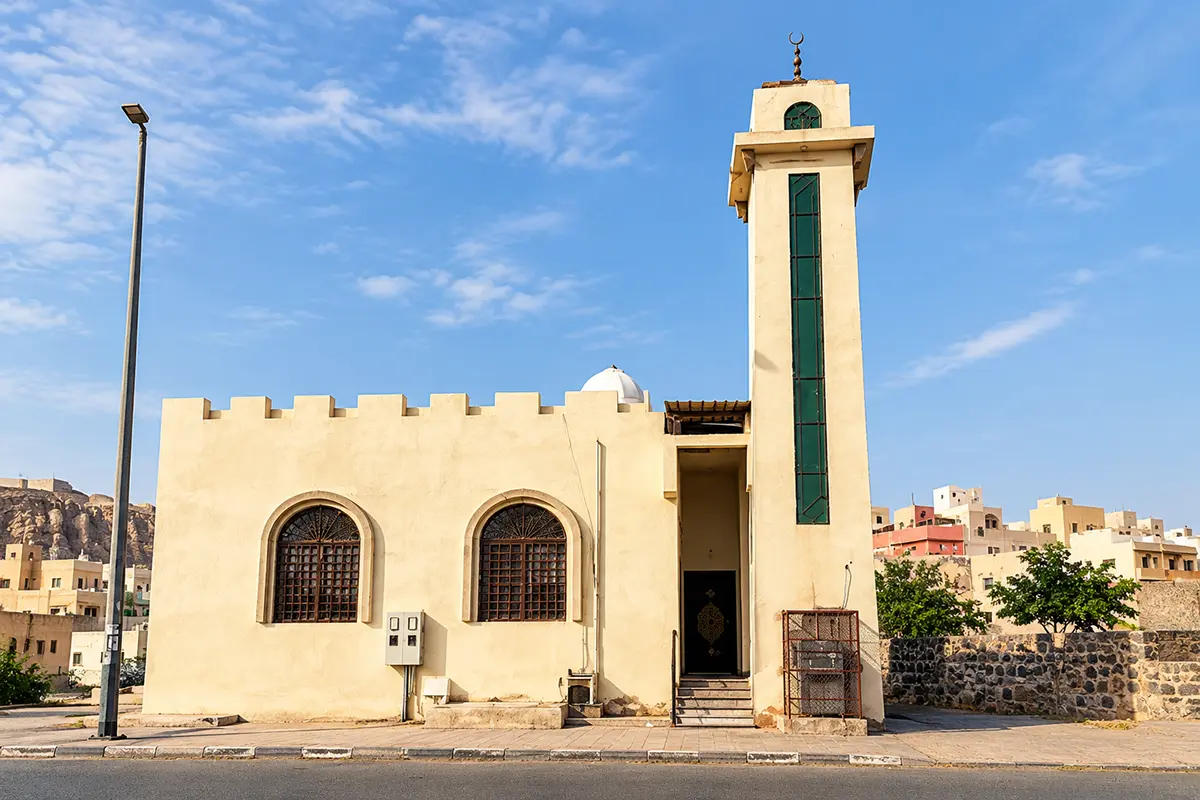 A small plain cream stone mosque with a low dome on a quiet street in the southern part of Madinah, seen in soft late afternoon light.
