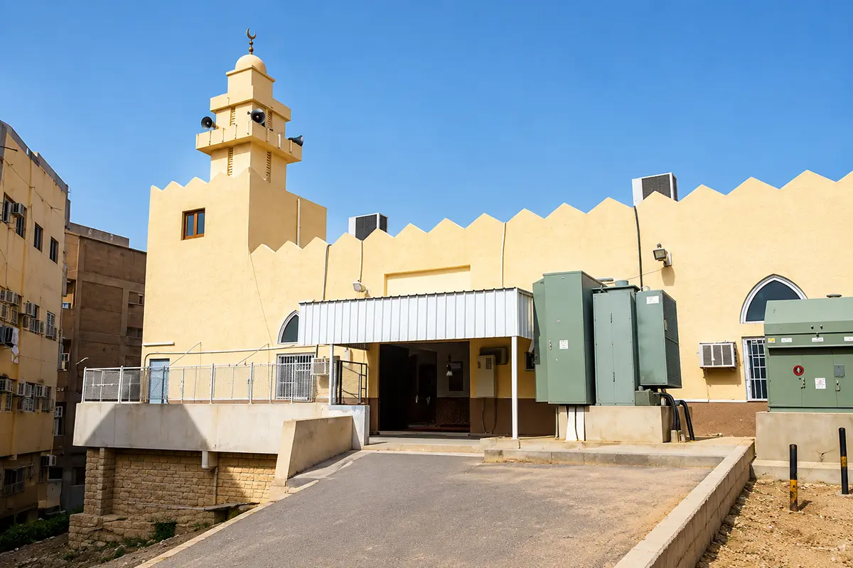 A small plain cream stone neighbourhood mosque with a low dome and short minaret on a quiet Madinah street, seen in warm late afternoon light.