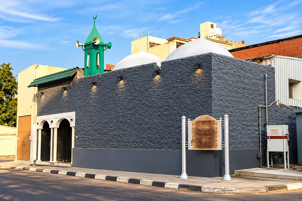 A small plain cream stone mosque beside the road north out of Madinah towards Uhud, with the red mass of the mountain faintly visible in the distance.