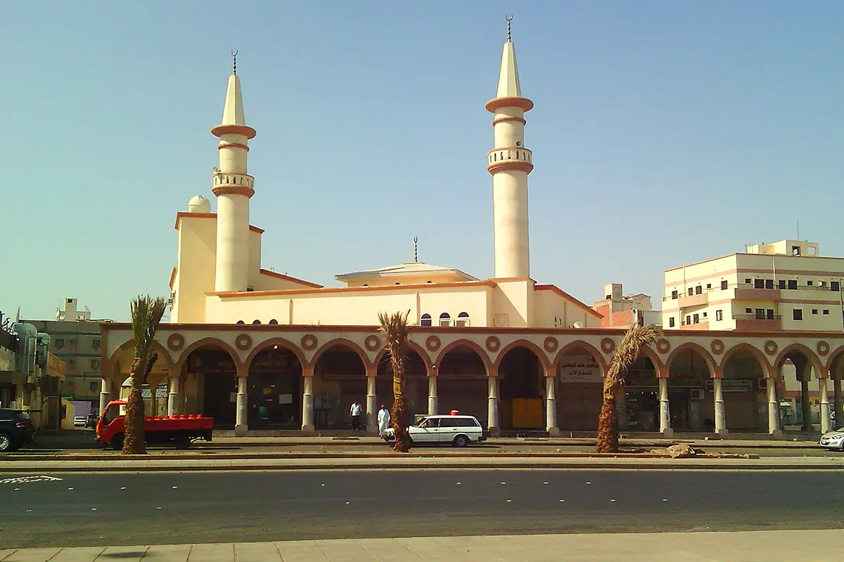 A modest modern cream stone mosque in Madinah with two slim minarets rising either side of a low dome, seen from the street in warm afternoon light.