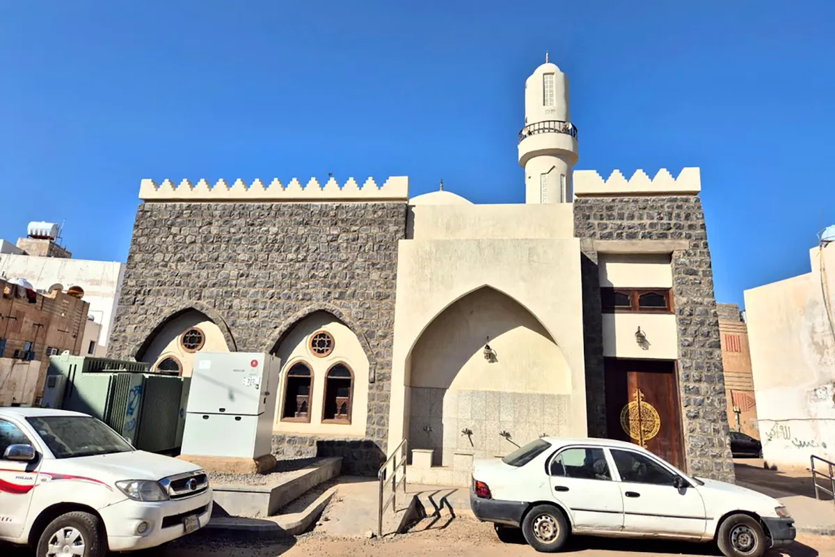 A small plain cream stone mosque in a quiet Madinah neighbourhood on the western side of Mount Sela', with the dark rocky ridge rising behind it in afternoon light.