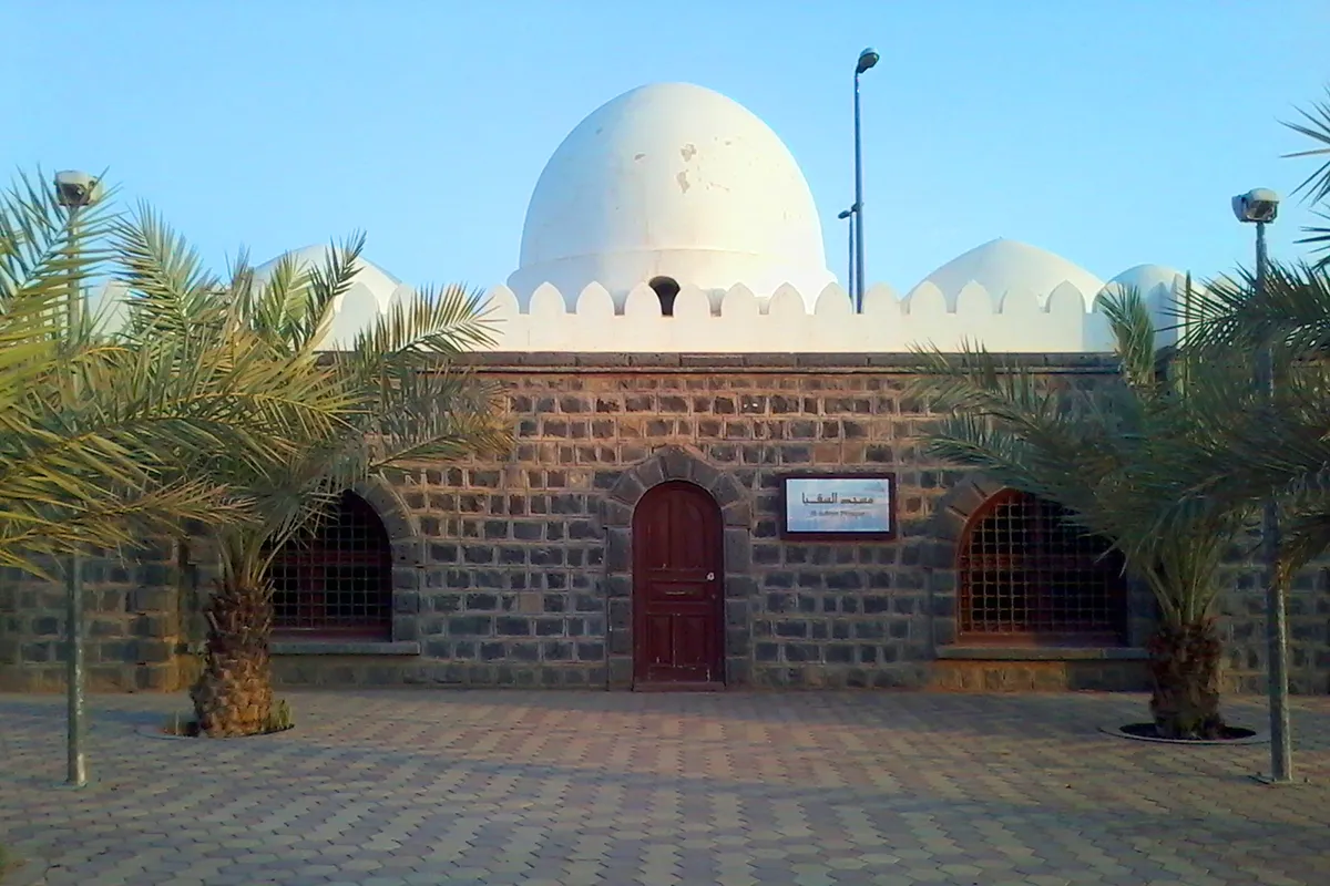 The small three-domed Masjid al-Suqya in the Anbariyah district of Madinah, with the dark stone arches of the old Hejaz Railway buildings behind it in afternoon light.