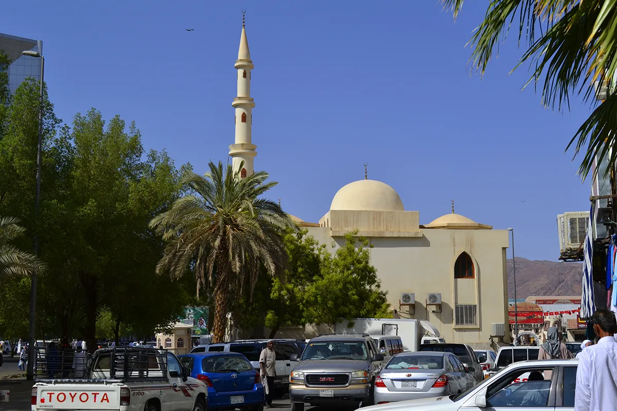 A very small plain cream stone mosque with a single slim corner minaret on a quiet street north of the Prophet's Mosque in Madinah, in warm afternoon light.
