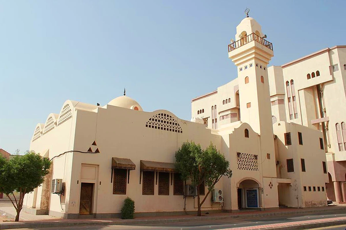 A small plain cream stone mosque raised slightly above the roadside on the route to Uhud in Madinah, with a low dome and short minaret in afternoon light.