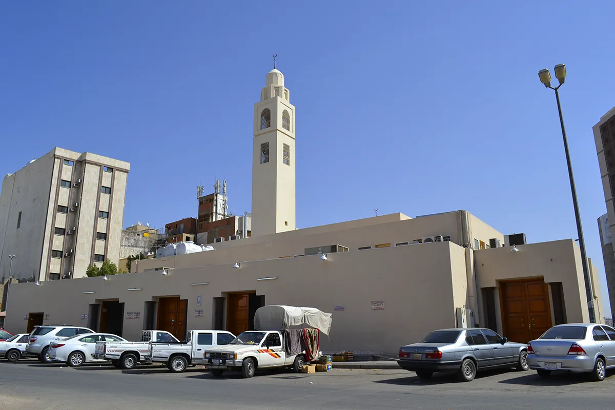 The plain cream stone facade and single slim minaret of Masjid al-Ijabah on a quiet Madinah street, seen in warm late afternoon light with no people in focus.