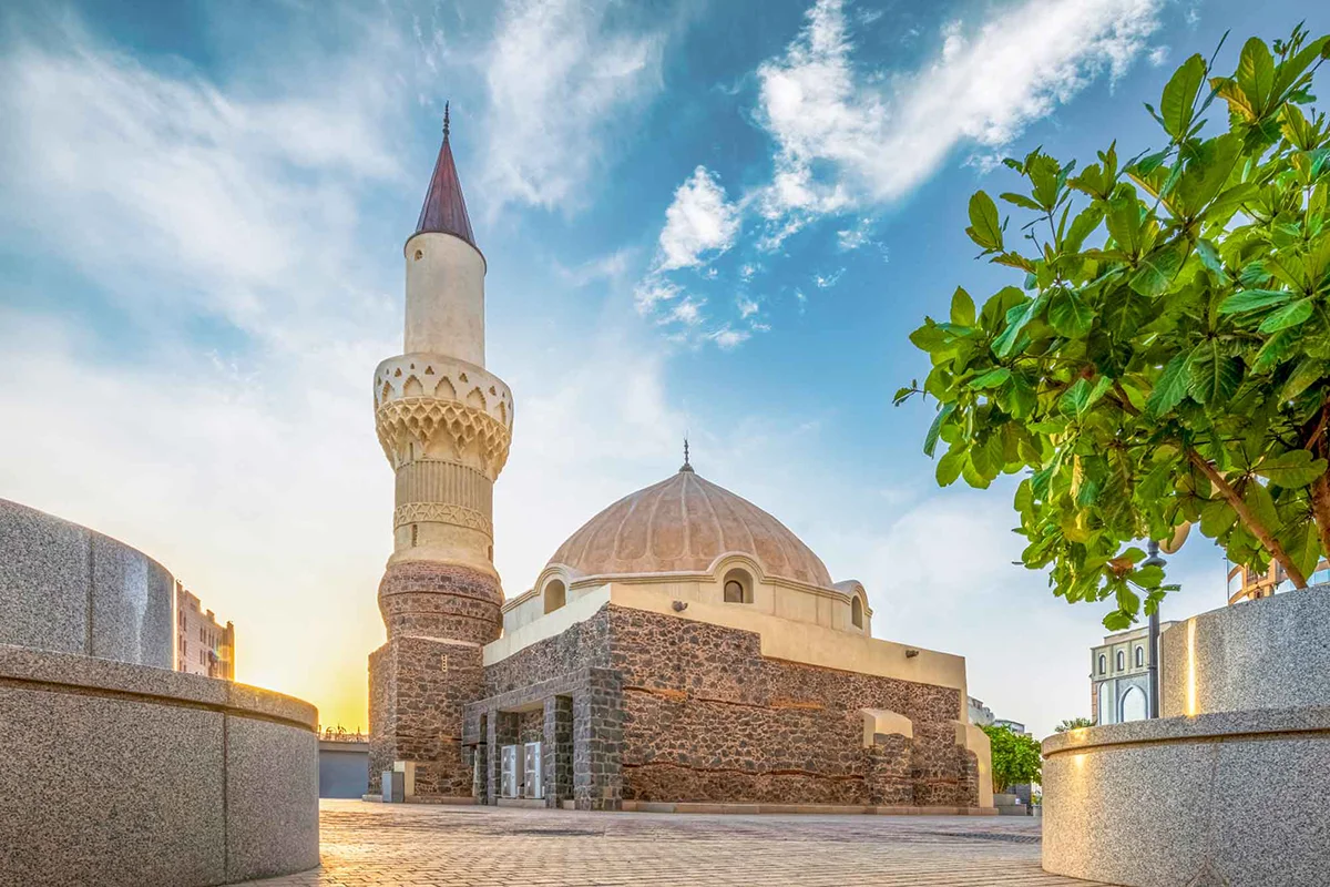The open rocky slope at the Seven Mosques site in Madinah where a small historic mosque once stood, with the remaining plain mosque buildings and Mount Sela' behind.