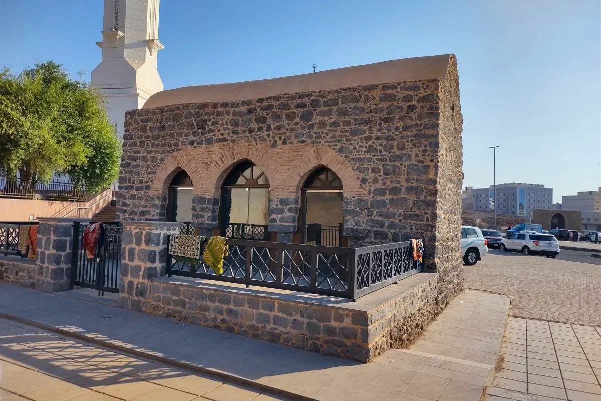 A small plain whitewashed historic mosque with a low vaulted roof standing against the bare rock at the foot of Mount Sela' in Madinah, in warm afternoon light.