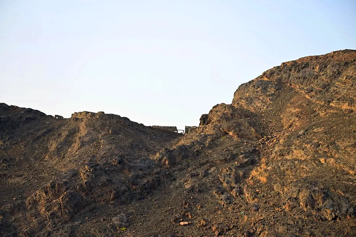 The long dark split ridge of Mount Sela' rising directly behind low modern buildings in western Madinah, its bare rock modelled by low late afternoon light.
