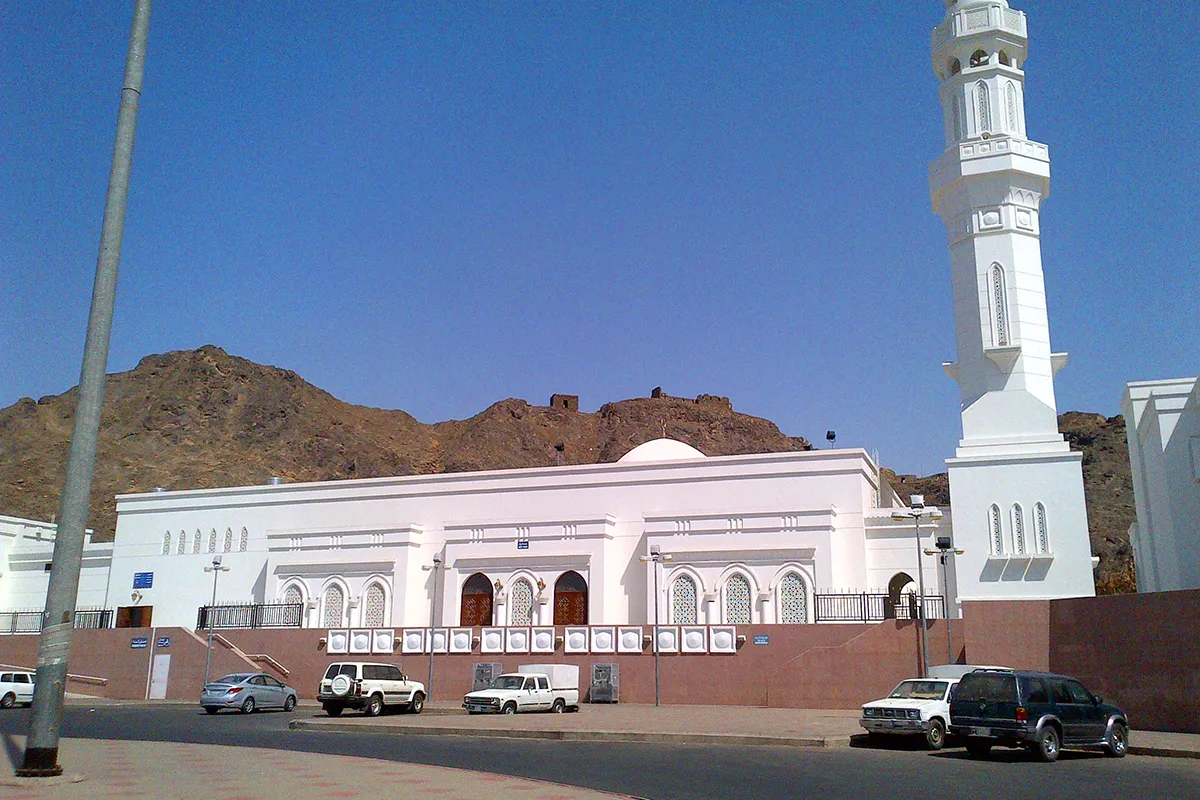 Small plain historic mosque buildings on the rocky lower slope of Mount Sela' in Madinah, with the dark ridge rising behind them in warm late afternoon light.