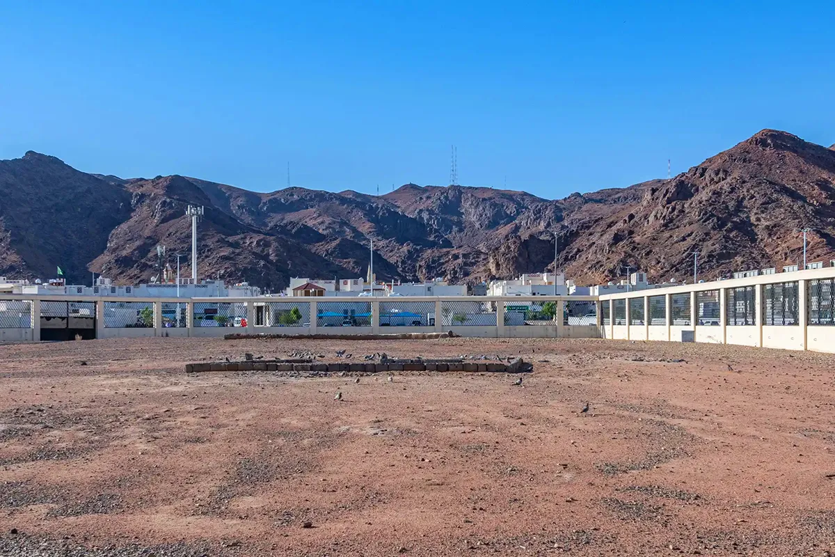 The low walled enclosure of the Martyrs of Uhud cemetery with plain unmarked graves inside a metal fence, the red rock of Mount Uhud rising directly behind in morning light.