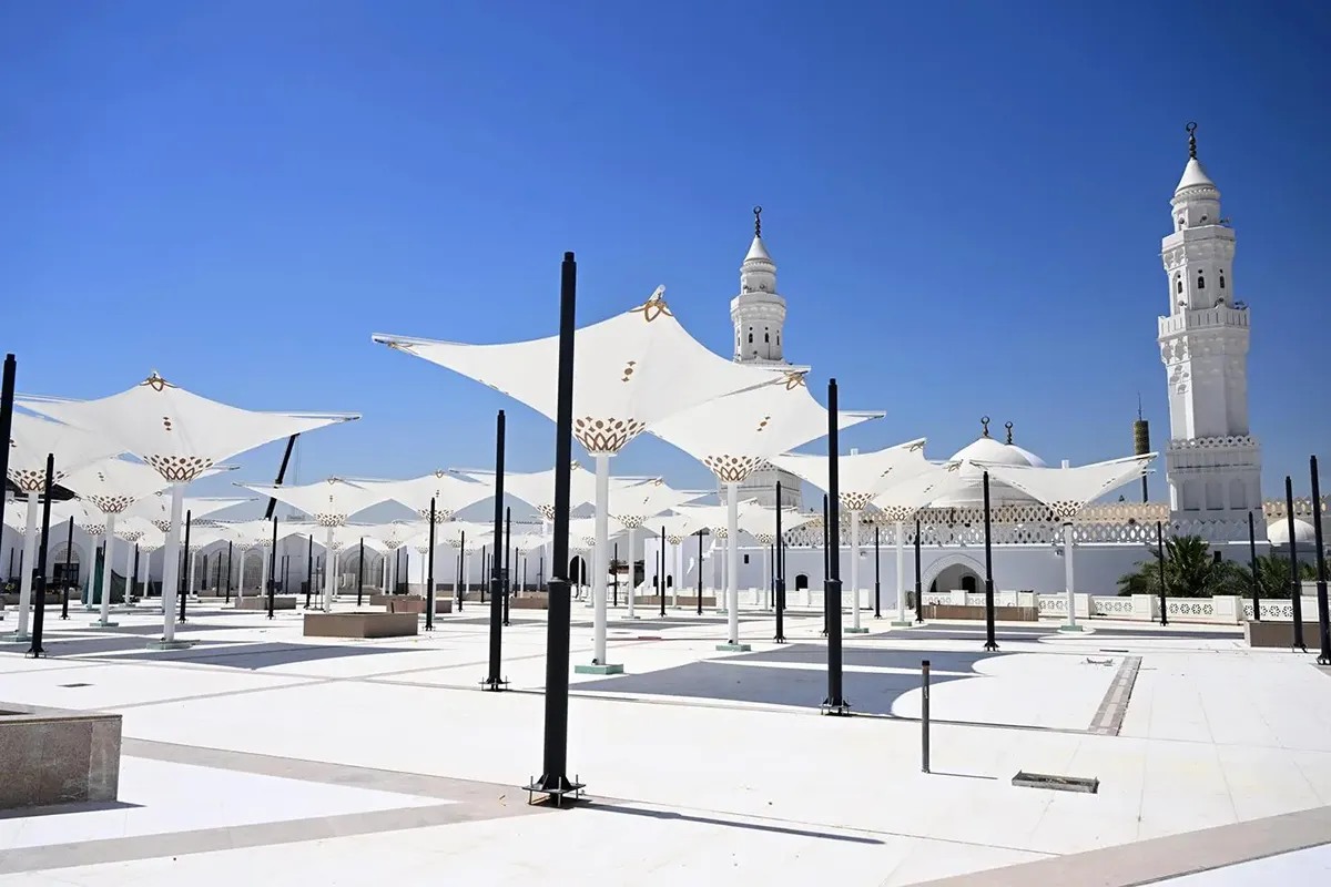 The white stone exterior of Masjid al-Qiblatayn in Madinah with its two domes and twin minarets against a clear sky, seen from the forecourt in warm late afternoon light.