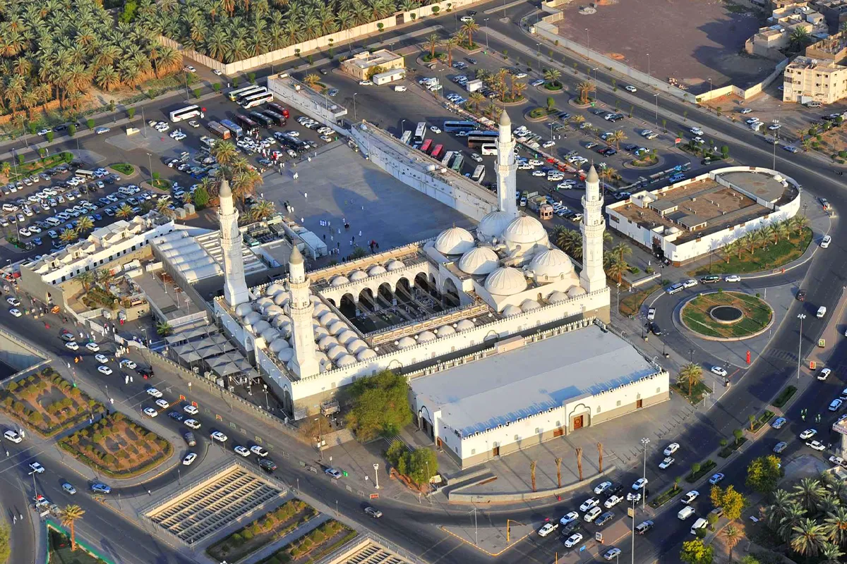 The white stone facade and slender minarets of Masjid Quba in Madinah under warm late afternoon light, with the wide paved forecourt in front and palm trees at the edge.