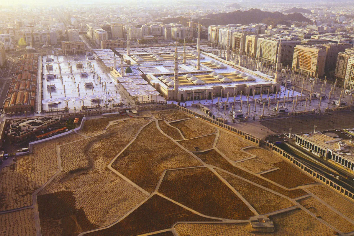 Rows of plain unmarked graves at Jannat al-Baqi in Madinah, each a low rectangle of bare earth edged with rough grey stones, with the minarets of the Prophet's Mosque behind in soft morning light.