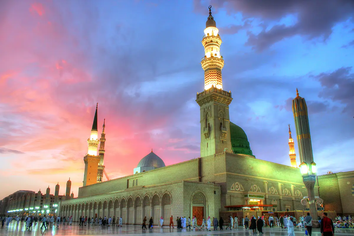 The courtyards of the Prophet's Mosque in Madinah at mid-morning with the large white retractable umbrellas open, minarets rising behind and worshippers crossing the marble as anonymous blurred shapes.