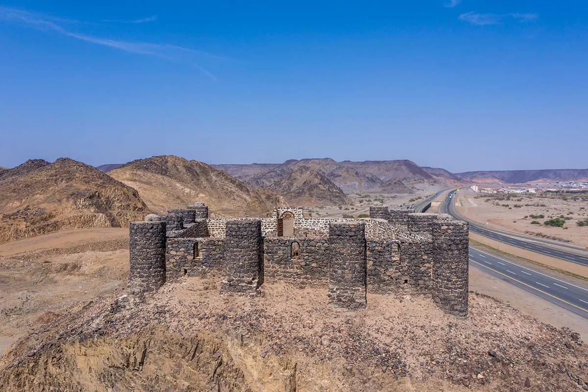 A small square stone fortress with corner towers standing on a low rocky rise at Asfan, dark basalt walls weathered and partly broken, flat gravel country stretching away behind it.