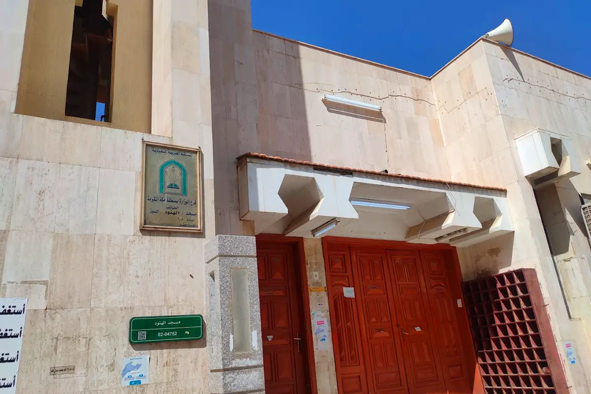 An old mosque in the central quarter of Taif seen from a narrow alley, plain rendered walls and a slender minaret above the rooftops, shopfronts and awnings crowding the lane below.