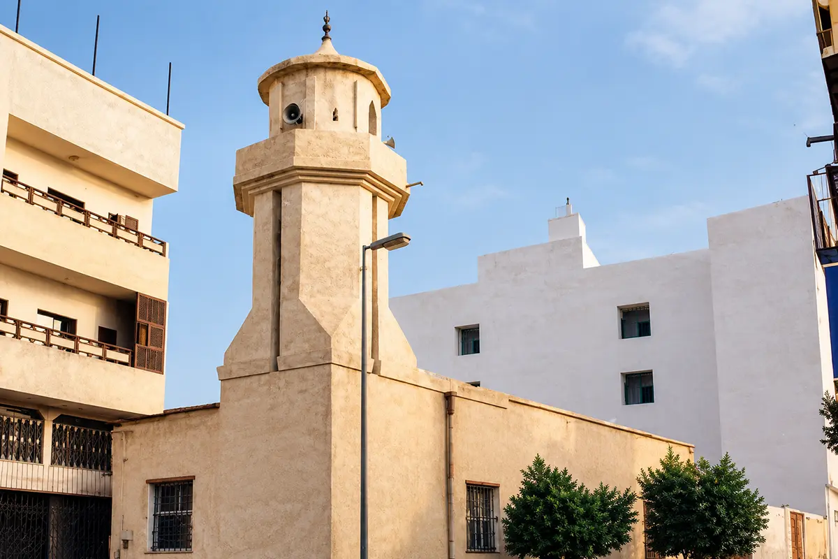 A small restored stone mosque in a quiet Taif residential street, thick pale walls with narrow windows, a short square minaret and modern houses standing close on either side.