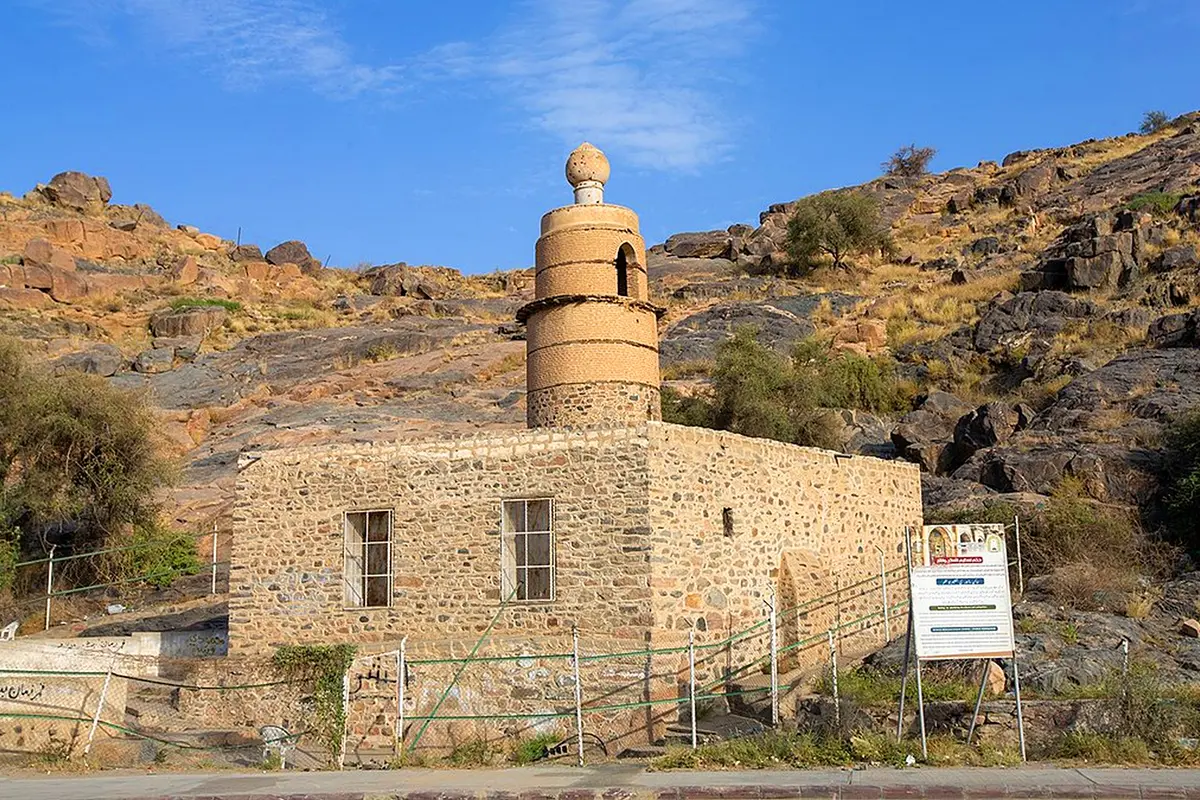 A small old stone mosque in a Taif farming village, a large natural boulder built into one corner of its wall, orchards and a dry watercourse behind it under a clear sky.
