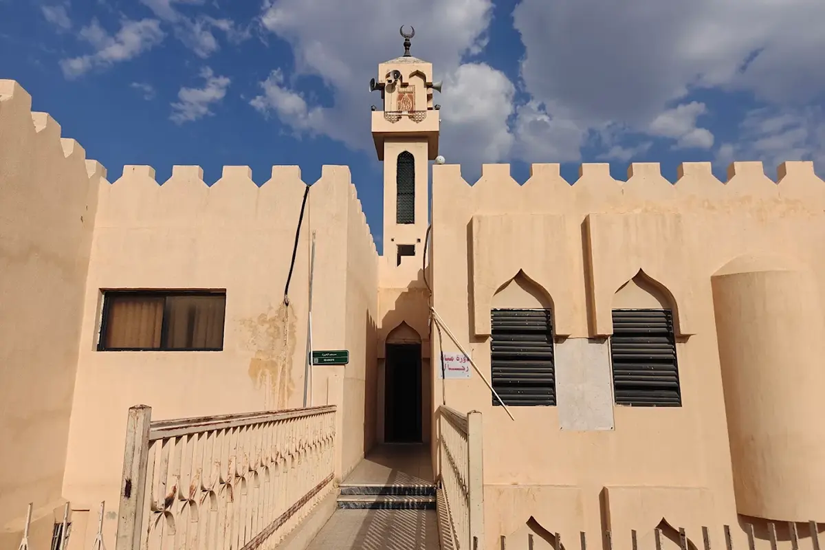An old restored stone mosque in the historic quarter of Taif, thick pale walls and a squat minaret, narrow alley shopfronts on either side and worn paving underfoot.
