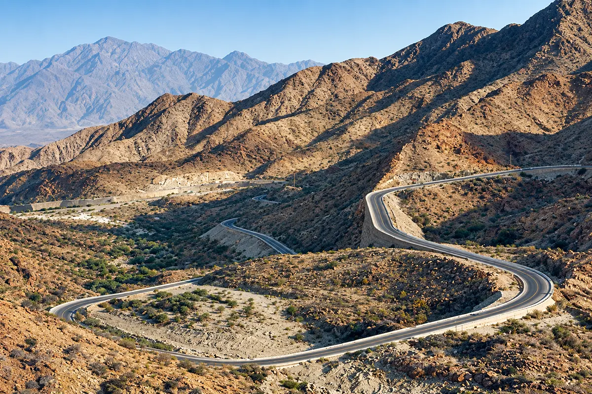 A wide dry valley between steep barren rock ridges on the road from Makkah towards Taif, pale gravel bed narrowing between the slopes, thin scrub and a modern highway running along one side.