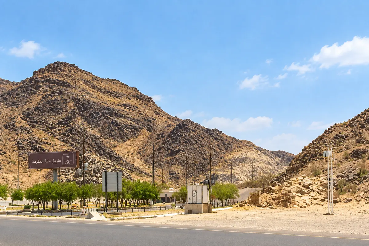 A narrow dry valley between Muzdalifah and Mina, pale gravel bed about twenty metres across running between low rocky banks, a modern road and pedestrian way carried above it.