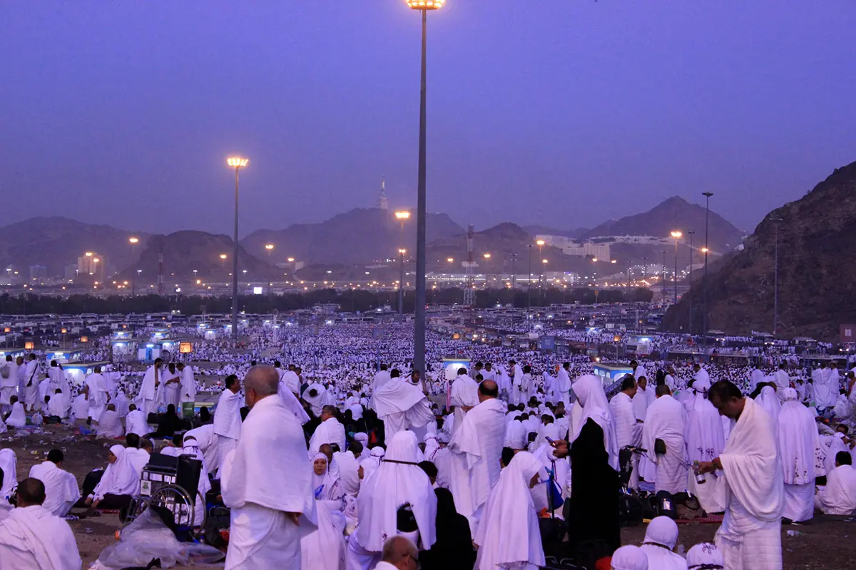 The open gravel ground of Muzdalifah at dusk, low bare ridges on either side, pilgrims lying and sitting in the open with no tents, sleeping mats spread directly on the stones.