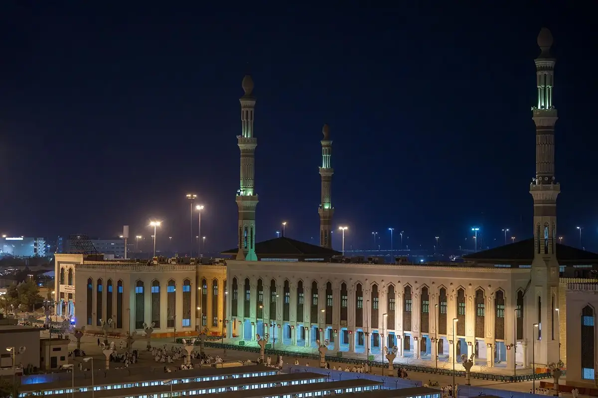The long low white mass of Masjid Namirah on the edge of the Arafat plain, six tall minarets rising above wide arched walls, bare brown hills behind and empty gravel ground in front.